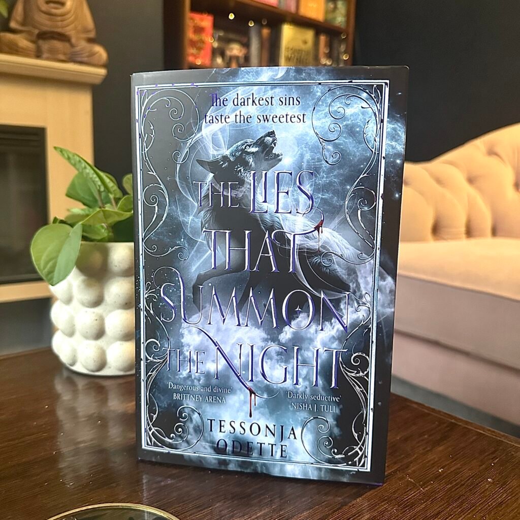 A hardback copy of The Lies That Summon The Night placed on a dark wood coffee table with a bookcase and pink sofa behind